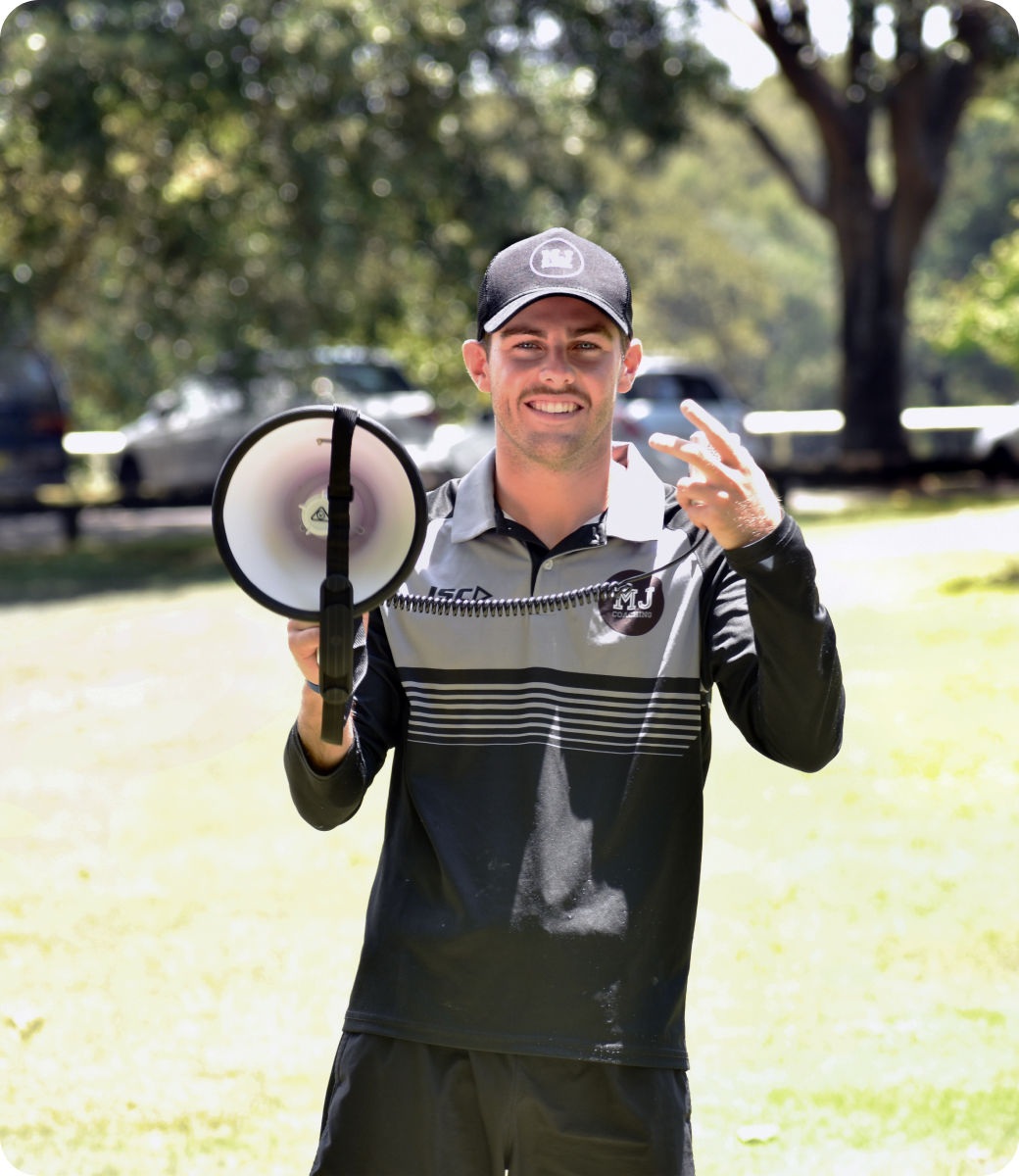 Edge founder Mac Jenkins standing outdoors in a park, smiling and holding a megaphone, wearing a cap and a black-and-gray athletic top.