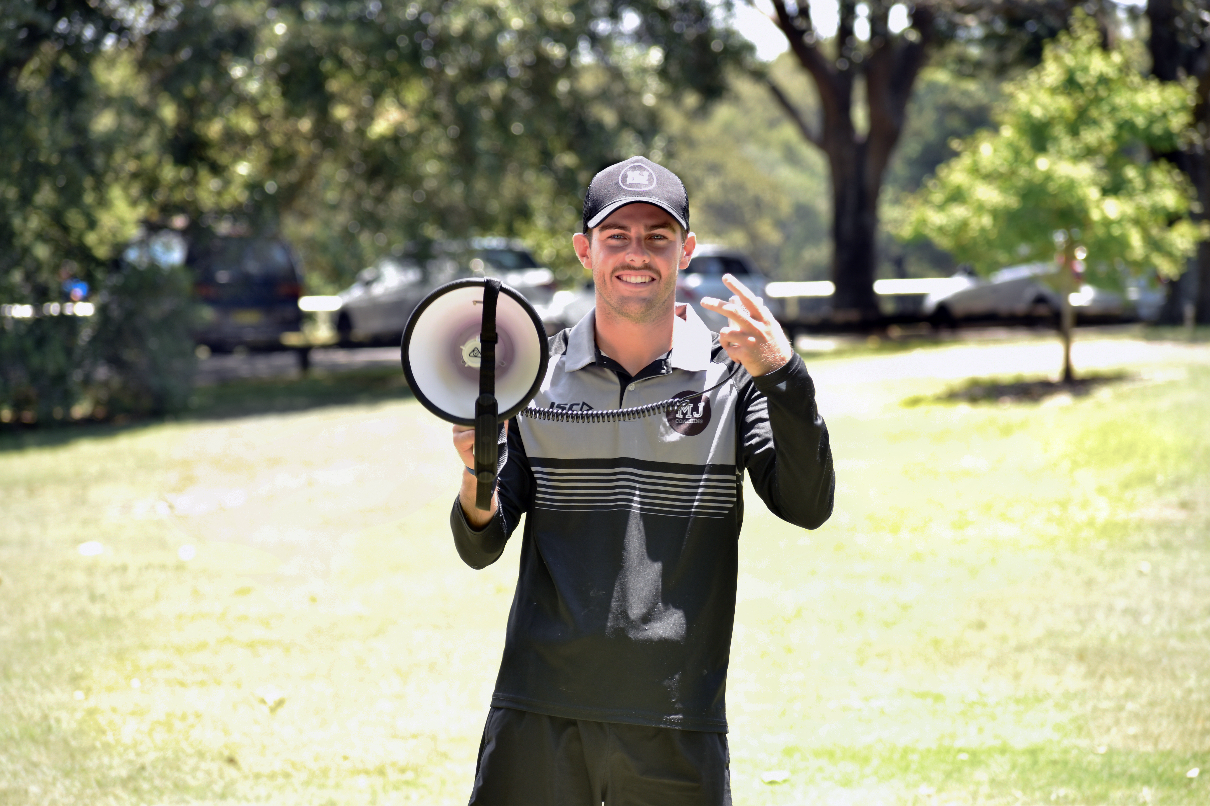 Edge founder Mac Jenkins standing outdoors in a park, smiling and holding a megaphone, wearing a cap and a black-and-gray athletic top.